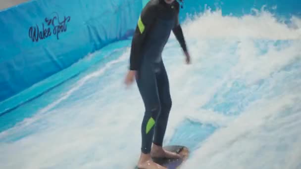 Adolescente está montando tabla de surf en simulador de olas en el interior. Joven surfista durante el entrenamiento en olas generadas. Actividad deportiva acuática. Surfer niño disfrutar del surf en interior imitador de surf. Entrenamiento de surf — Vídeos de Stock