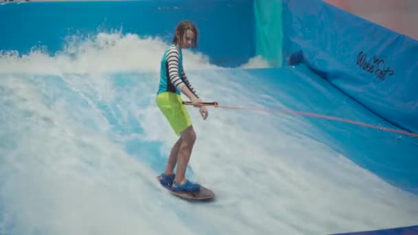 Adolescente está montando prancha de surf no simulador de ondas dentro de casa. Surfista jovem durante o treino em ondas geradas. Actividades desportivas aquáticas. Surfista garoto gosta de surfar no imitador de surf indoor. Treinamento de surf — Vídeo de Stock