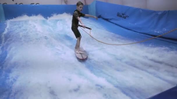 Teenager rider surf board på bølge simulator indendørs. Unge surfer under træning på genererede bølger. Vandsportsaktiviteter. Surfer kid nyde surfing på indendørs surf imitator. Surfing uddannelse – Stock-video