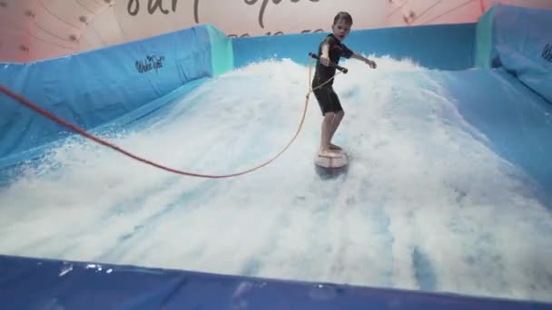 Adolescente está montando prancha de surf no simulador de ondas dentro de casa. Surfista jovem durante o treino em ondas geradas. Actividades desportivas aquáticas. Surfista garoto gosta de surfar no imitador de surf indoor. Treinamento de surf — Vídeo de Stock