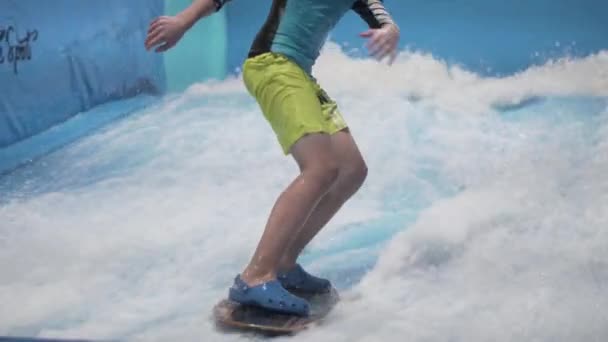 Nastolatek jedzie na desce surfingowej na symulatorze fal w domu. Młody surfer podczas treningu na generowanych falach. Sporty wodne. Dzieciak surfera lubi surfować na imitatorze krytego surfingu. Trening surfingu — Wideo stockowe