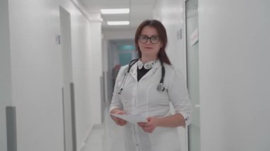 Kadın doktor hastane koridorunda duruyor. Modern bir klinikte poz veren gözlüklü ve beyaz önlüklü genç bir kadın portresi. Gururlu profesyonel kadın doktor kameraya bakıyor.