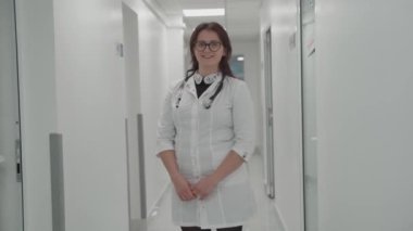 Tıbbi enstitüde duran kendine güvenen genç bayan doktor. Gözlüklü profesyonel doktor kadın hastane koridorundaki kameraya bakıyor. Pratisyen hekimin portresi