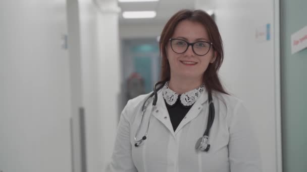 Portrét usmívající se tisícileté doktorky v lékařské uniformě a stetoskopu, jak se dívá na kameru na chodbě moderní nemocnice. Zdravotní koncepce, zdravotní pojištění — Stock video
