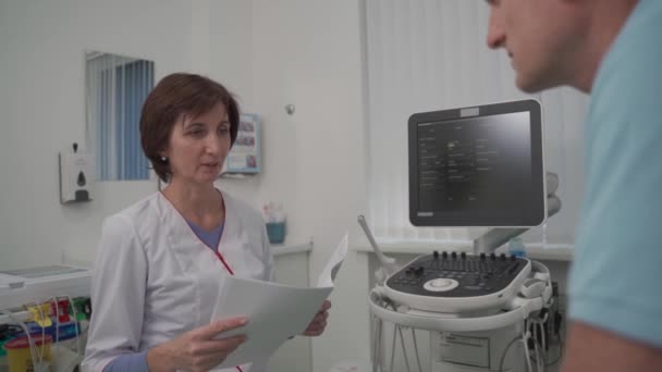 Tema de salud y medicina. Médico general femenino verá pruebas médicas durante el examen de salud del paciente masculino en el consultorio de la clínica. Trabajador médico en sala de diagnóstico por ultrasonido con paciente — Vídeo de stock