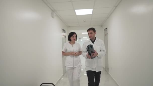 Два доктора идут по коридору и обсуждают МРТ мозга. Медицинский работник, выглядящий как рентген пациентов, идущих по коридору клиники. Профессия, люди, здравоохранение и медицина — стоковое видео
