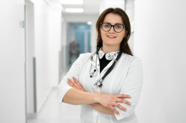 Kadın doktor hastane koridorunda duruyor. Modern bir klinikte poz veren gözlüklü ve beyaz önlüklü genç bir kadın portresi. Gururlu profesyonel kadın doktor kameraya bakıyor.