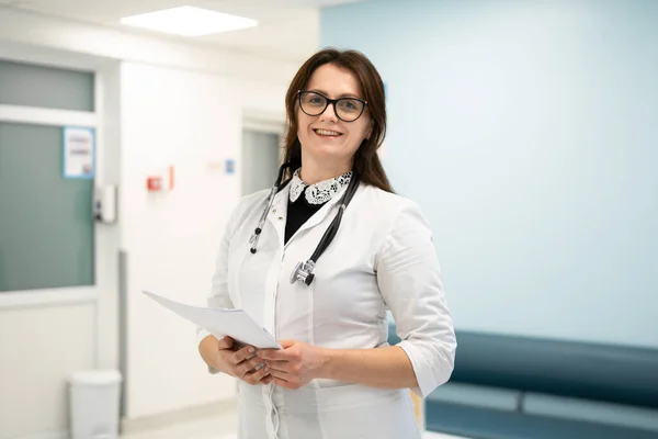 Ritratto di una sorridente dottoressa millenaria in uniforme medica e stetoscopio che guarda la macchina fotografica nel corridoio di un moderno ospedale. Concetto di assistenza sanitaria, assicurazione medica — Foto Stock