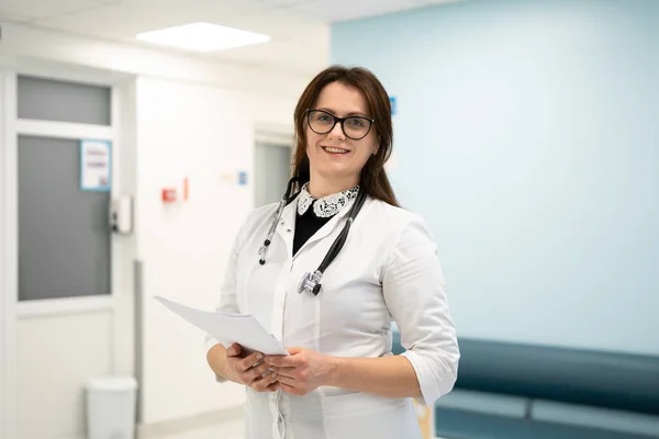Жінка-лікар стоїть в лікарняному коридорі. Портрет молодої жінки-лікаря в окулярах і біле пальто, що позує в сучасній клініці. Горда професійна жінка-лікар терапевт дивиться на камеру — стокове фото