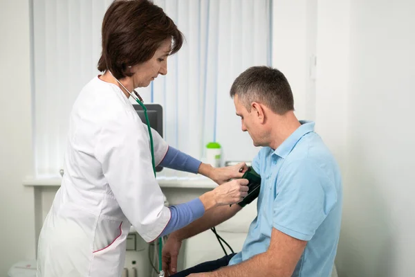 La doctora mide la presión arterial con un esfigmomanómetro al paciente masculino en el examen médico en el consultorio de la clínica. Cardiólogo comprueba la presión arterial del paciente masculino en el hospital de cardiología — Foto de Stock