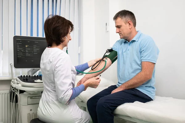 Il medico femminile misura la pressione sanguigna con uno sfigmomanometro al paziente di sesso maschile all'esame medico in ambulatorio. Cardiologo controlla la pressione sanguigna del paziente maschile presso l'ospedale di cardiologia — Foto Stock