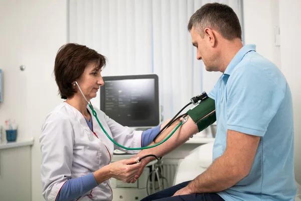 La doctora mide la presión arterial con un esfigmomanómetro al paciente masculino en el examen médico en el consultorio de la clínica. Cardiólogo comprueba la presión arterial del paciente masculino en el hospital de cardiología — Foto de Stock