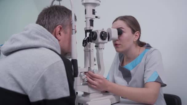 眼科医と患者の検査視力。男は観光客と目のテストを行う。眼科医は、診療所で目の検査のための機器を使用しています。視力測定を行う患者を調べる医師 — ストック動画