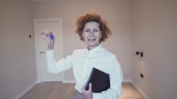 幸せな高齢女性の不動産業者は、家賃のための空のアパートの背景にカメラへの不動産への鍵を示しています。成熟した不動産屋は取引に満足しています。賃貸アパート — ストック動画