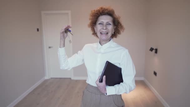 Felice donna anziana agente immobiliare mostra le chiavi del bene immobile per la fotocamera sullo sfondo di un appartamento vuoto in affitto. Agente immobiliare maturo felice con l'affare. Appartamento in affitto — Video Stock