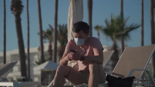 Summer vacation concept. Epidemic coronavirus covid-19 flu virus. New normal in vacation in time after COVID-19 pandemic. Sad masked man sits on sun lounger on the territory of an empty hotel — Stockvideo