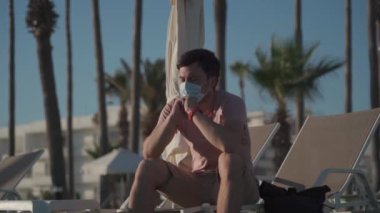 Salgın sırasında turizm. İyi yolculuklar. Coronavirus seyahat yasağı. Koronavirüs salgını nedeniyle karantina. Hasta, maskeli genç bir adam boş bir otelin bölgesinde güverte sandalyesinde oturuyor.