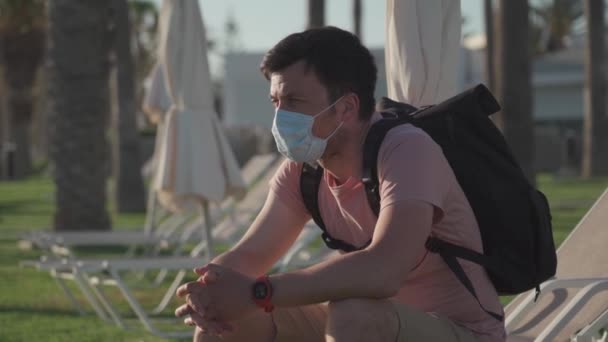 Triste hombre con máscara protectora se sienta en una tumbona en un hotel vacío en Chipre durante la cuarentena y covid 19 pandemia. Turismo durante la pandemia. Buen viaje. Prohibición de viajar por el Coronavirus. Epidemia de Coronavirus — Vídeos de Stock