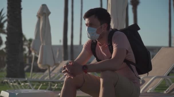 Triste hombre con máscara protectora se sienta en una tumbona en un hotel vacío en Chipre durante la cuarentena y covid 19 pandemia. Turismo durante la pandemia. Buen viaje. Prohibición de viajar por el Coronavirus. Epidemia de Coronavirus — Vídeos de Stock