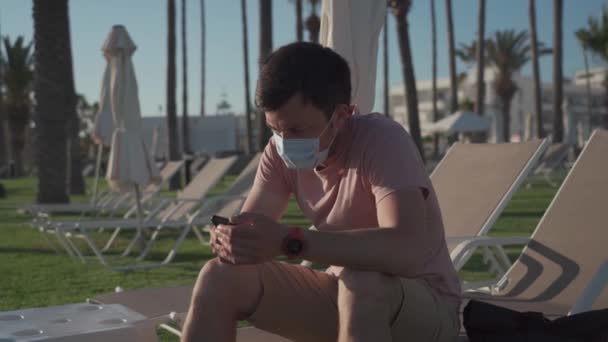 Tourism during pandemic. Safe traveling. Coronavirus travel ban. Quarantine due coronavirus epidemic. Sick tired young man in face mask sit on deck chair on the territory of an empty hotel — стокове відео