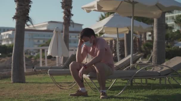 Summer vacation concept. Epidemic coronavirus covid-19 flu virus. New normal in vacation in time after COVID-19 pandemic. Sad masked man sits on sun lounger on the territory of an empty hotel — Stock Video