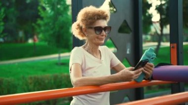 Sportif olgun bir kadın yoga dersinden sonra sokak spor salonunda telefon kullanıyor. Üst düzey Kafkasyalı bayan dışarıda yoga antrenmanından sonra mola veriyor. Aktif yaşam tarzı konsepti. İnsanlar, spor ve teknoloji