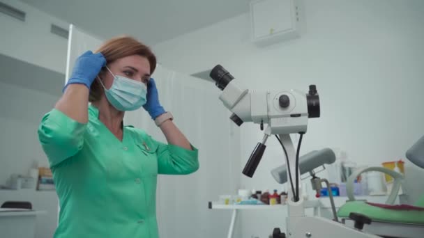 Πρόληψη και διάγνωση του καρκίνου του τραχήλου. Ώριμη γυναίκα γιατρός που βάζει μια προστατευτική μάσκα πριν από την εργασία με ένα μικροσκόπιο colposcopy σε ένα ginigological γραφείο μιας σύγχρονης γυναικείας κλινικής υγείας — Αρχείο Βίντεο