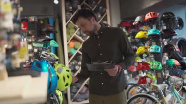 Bisikletçi bir erkek, bisiklet dükkanında kask envanteri çıkarıyor. Elinde bir pano olan bir spor mağazasının sahibi vitrindeki bisiklet kasklarının fiyatlarını kontrol eder.
