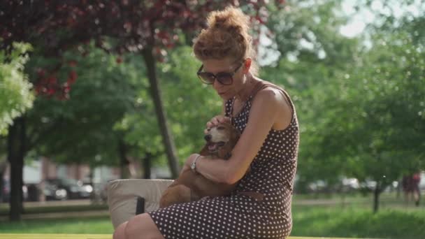 Dojrzała kobieta siedzi na ławce w parku miejskim z jamnikiem trzymającym ją w ramionach i przytulającym. Szczęśliwa staruszka siedząca z psem na ławce w letnim parku. Reszta ze zwierzątkiem — Wideo stockowe