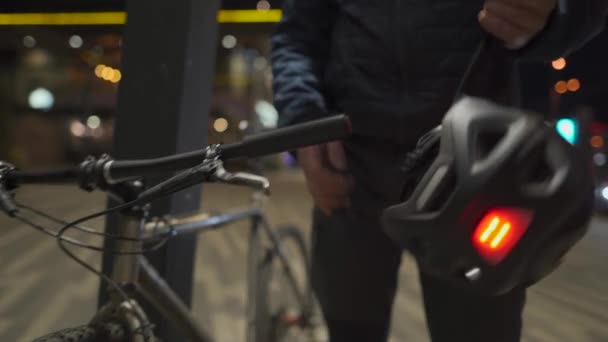 Мужчина-велосипедист в медицинской маске надевает защитный шлем с мигающим светом на задней части шлема и забирает велосипед с парковки вечером в городе. Пригородный велосипед для концепции covid-19 — стоковое видео
