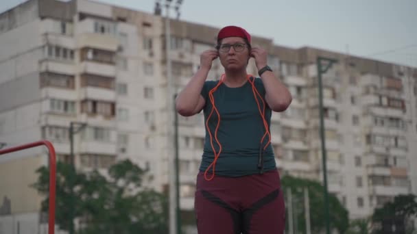 Wanita Kaukasia yang kelebihan berat memakai headphone dan menyalakan musik dengan jam tangan pintar sebelum berlatih lompat tali di stadion kota pada musim panas. Tema melenceng, tujuan menurunkan berat badan. Olahraga dan kesehatan — Stok Video