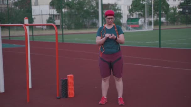 Tema de esportes, música em fones de ouvido, treino cardiovascular com sobrepeso e objetivo de perda de peso. Mulher com excesso de peso coloca fones de ouvido e liga a música antes de treinar pulando corda no estádio da cidade no verão — Vídeo de Stock