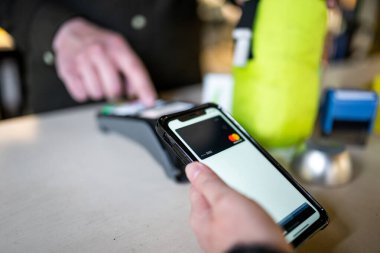 Kasiyer elinde NFC terminalini tutarken modern akıllı telefonu kullanarak kasaya para ödeyen bir kadın görüntüsü. Akıllı telefon ve NFC teknolojisi kullanılarak kablosuz ödeme.