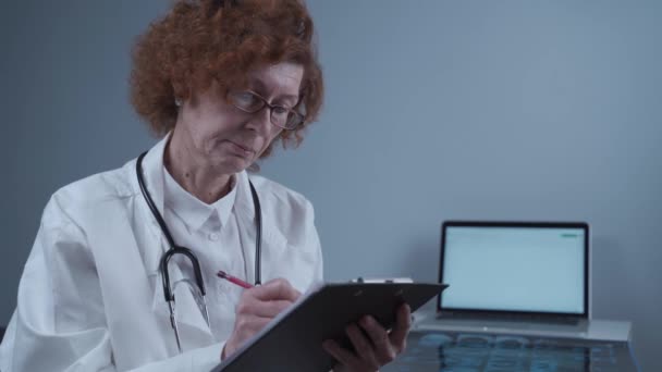Концентрированная женщина-радинолог пишет записи в регистрационную книгу, работает в клинике. Квалифицированный профессиональный пожилой врач, назначающий лечение заболеваний, занимающийся бумажной работой — стоковое видео