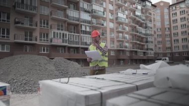 Miğferli ve giyimli yenilikçi bir müfettiş malzemeleri kontrol eder ve bir konut binasının inşaat alanındaki belgelerle karşılaştırır. İnşaat müfettişi çalışıyor.