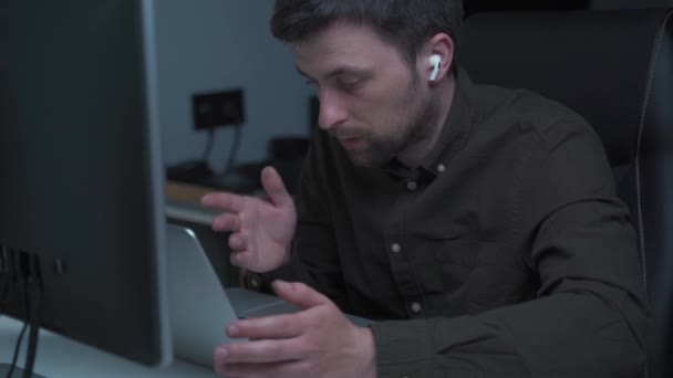 Mužský programátor pracuje u počítače a používá sluchátka v uchu k hovoru s kolegou nebo zákazníkem. Bezdrátový bílý sluchátko sluchátko v uchu muže IT vývojář v práci — Stock video