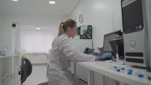 科学者の女性は研究室の彼女の職場で働き、パーソナルコンピュータを使用する。開発、分析、遺伝学。医薬品研究センター。微生物学者は研究室でコンピュータ上のデータをチェックします — ストック動画