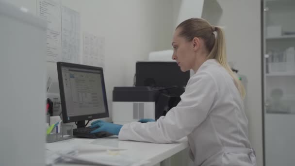 Modern klinikteki tıp laboratuarında çalışan kadın patoloji teknisyeni. Test sonuçları. Ciddi bir kadın hastanede çalışırken monitöre ve kan testlerine bakıyor. — Stok video