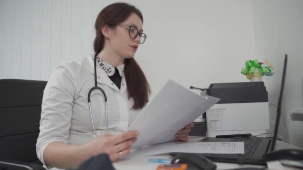 Una joven doctora en gafas se reúne con una paciente virtualmente usando tecnología de videollamadas, sostiene los resultados de las pruebas de los pacientes en sus manos y da consejos de tratamiento en una computadora portátil en el consultorio de la clínica — Vídeo de stock