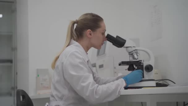 Γυναίκα επιστήμονας που χρησιμοποιεί μικροσκόπιο ενώ εργάζεται στο εργαστήριο. Εργαστήριο σύγχρονης ιατρικής έρευνας. Γυναίκα βοηθός εργαστηρίου ψάχνει στο μικροσκόπιο ανάλυση δειγμάτων βιοχημικές ουσίες, εξετάσεις αίματος — Αρχείο Βίντεο