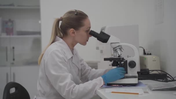 Kvinnlig läkare arbetar med prover och analyser i medicinskt laboratorium bakom mikroskop. Kvinnlig laboratorietekniker som analyserar prover genom mikroskop på sjukhus. Medicin, mikrobiologi — Stockvideo