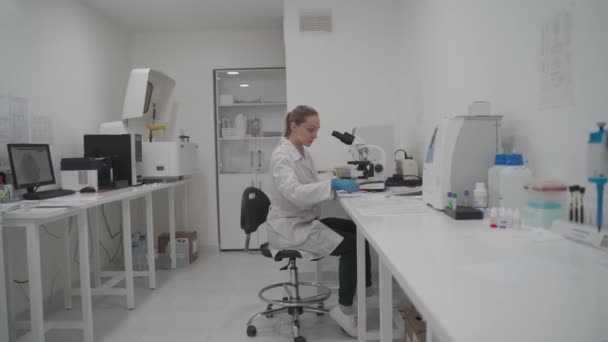 Médico femenino en uniforme trabajando con microscopio haciendo análisis en la oficina de laboratorio. Científico trabajando en laboratorio. Microbióloga mujer trabajando en laboratorio médico moderno con equipo tecnológico — Vídeos de Stock