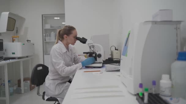 Profesional médico femenino trabaja con muestras y analiza en laboratorio médico detrás del microscopio. Técnico de laboratorio mujer analizando muestra a través del microscopio en el hospital. Medicina, microbiología — Vídeo de stock