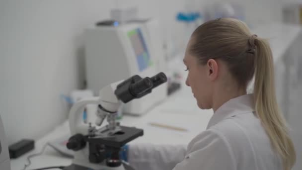 Vrouwelijke arts in uniform werken met microscoop het maken van analyse op laboratoriumkantoor. Wetenschapper werkt in het laboratorium. Microbioloog vrouw die werkt in een modern medisch lab met technologische apparatuur — Stockvideo