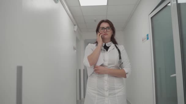 여성 일반 개업 의사는 병원 복도를 걷는 동안 환자에게 휴대 전화로 의사 상담을 하고, 서류나 의료 기록을 분석하며, 원격으로 조언을 해준다 — 비디오