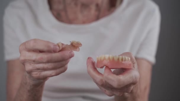 Donna anziana che tiene in mano il primo piano della protesi dentale. La vecchia mostra denti falsi. Dentiera rimovibile nelle mani di un paziente anziano in studio su sfondo grigio. — Video Stock
