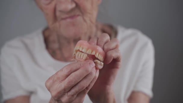 Mujer mayor sin dientes sostiene la dentadura postiza en sus manos y hace un movimiento de masticación divertido con sus dientes. Una vieja paciente ortodoncista muestra dientes postizos de cerca. Prótesis dental aplaude dientes — Vídeo de stock