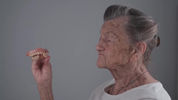 グレーの背景にスタジオで90歳の白髪の満足したシニア女性の手の中に歯科矯正。歯のないシニア女性保持していますデュエンスで彼女の手に笑顔 — ストック動画