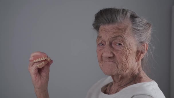 Wanita tua melepas gigi palsu dilepas, menunjukkan ke kamera dan tersenyum. Konsep perawatan lisan, gigi palsu dan usia tua. Dentur lengkap di tangan perempuan tua dengan rambut abu-abu studio video syuting — Stok Video