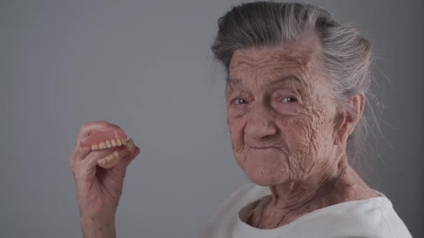 Знімний зуб в руках старшого пацієнта в студії на сірому фоні. Тема ортодонтії. Бабуся використовує зубний протез. Гнучкий нейлоновий протез. Повний стоматологічний тримач задоволений старшою жінкою — стокове відео